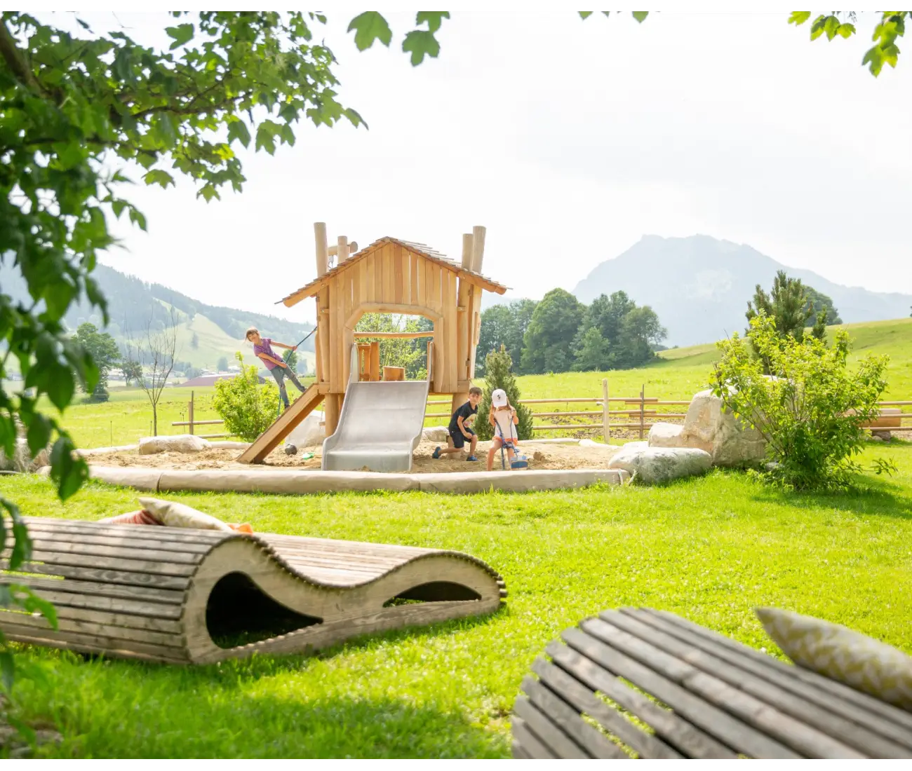 Holzliege auf Rasen vor Spielplatz und Bergkulisse